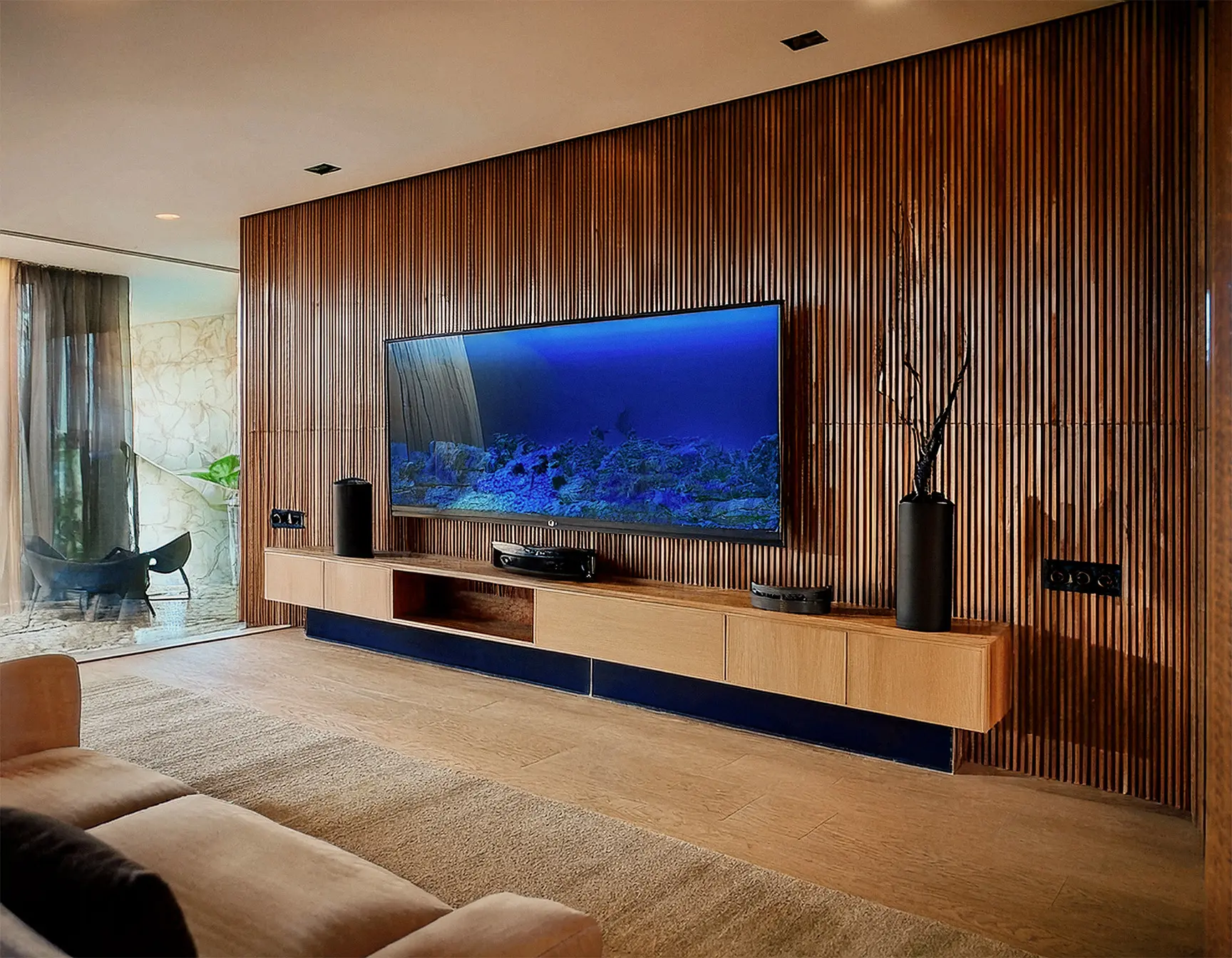 Uma sala moderna e com móveis de alto padrão, minimalistas e nota-se sua qualidade. A parede possuí detalhes em madeira ripada e nela está uma televisão segurada por um suporte. Abaixo dela ve-se um nicho suspenso com gavetas e cor off white dando todo detalhe e tom de sofisticação. Ao fundo também é possível ver uma área externa com cadeiras e uma vegetação